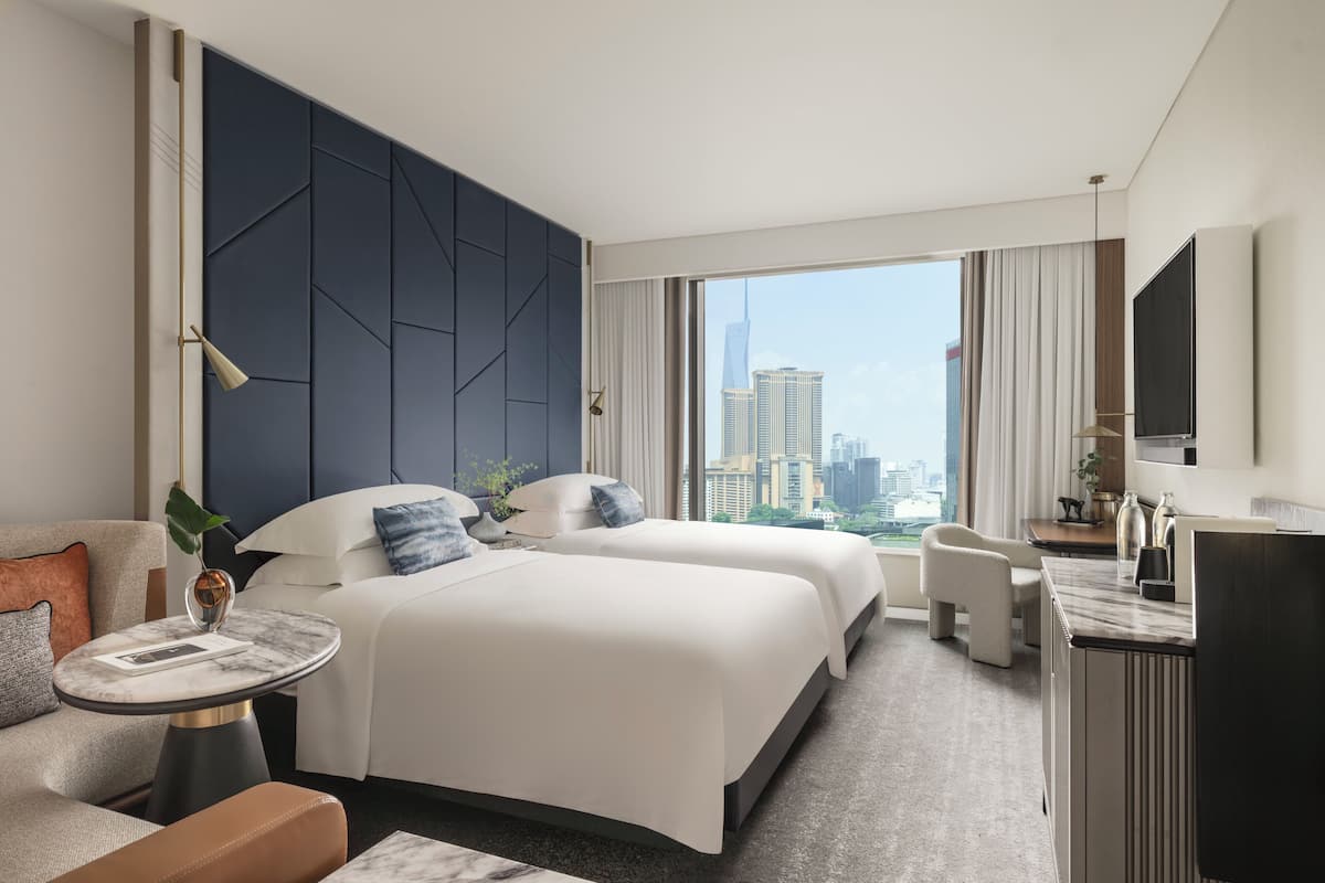 A modern hotel room with two beds, a desk, a chair, and a large window offering a city view.