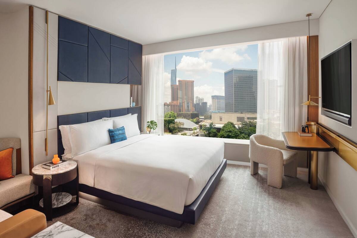 A modern hotel room with a large bed, a desk, a chair, a TV, and a city view.