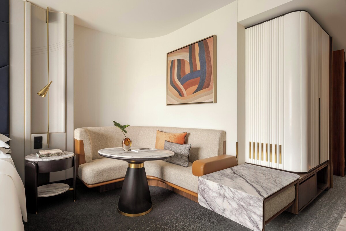 A modern hotel room with a sofa, a small round table, a cabinet, and a large abstract painting.