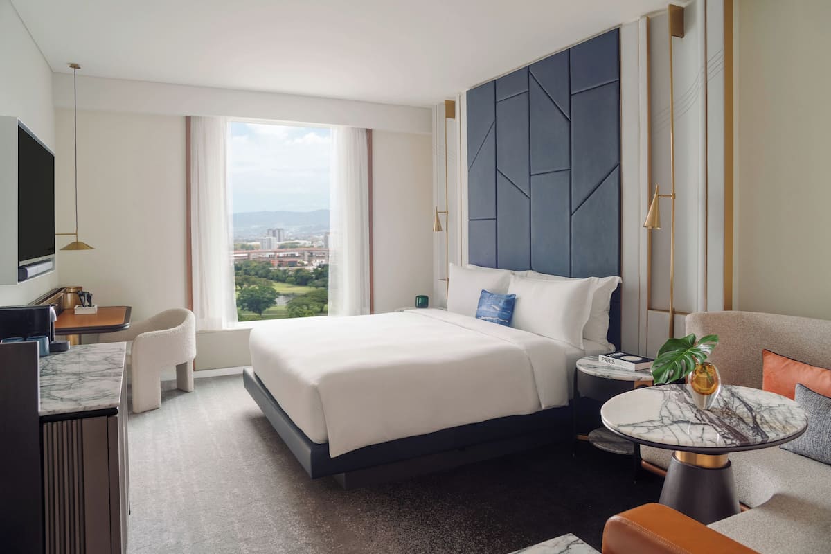 A hotel room with a large bed, a desk, a TV, a round table, and a view of the city.