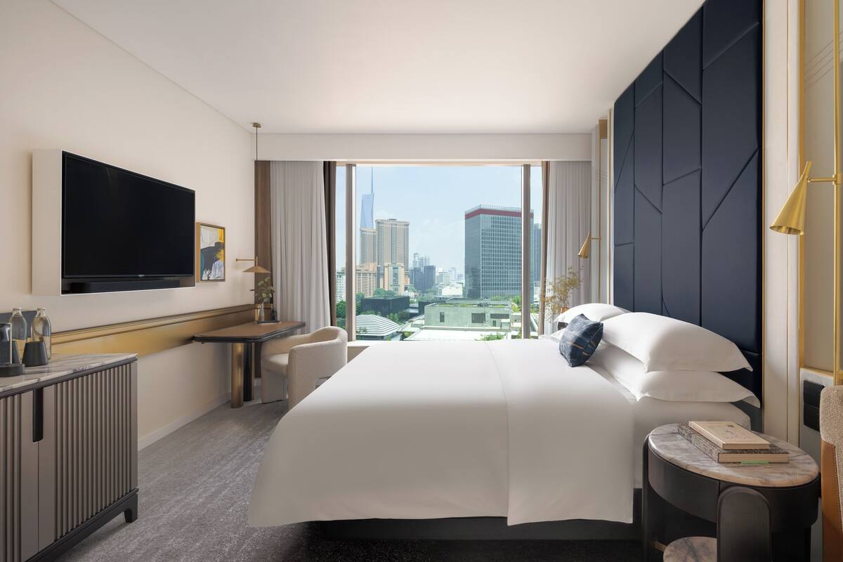 A hotel room with a large bed, a flat-screen TV, a desk, and a view of the cityscape.