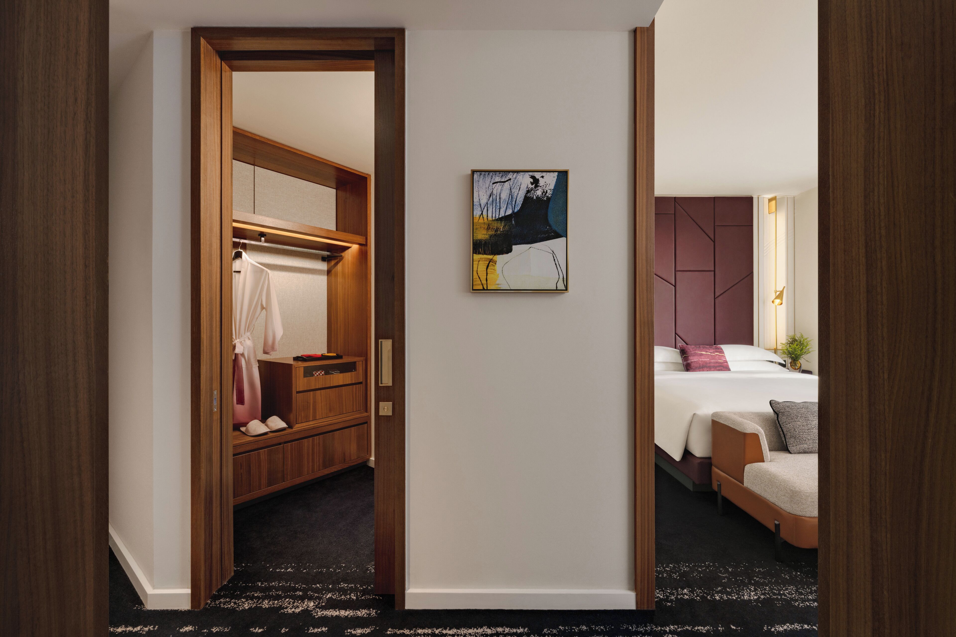 A hotel room with a wooden wardrobe, a bed with a headboard, a painting on the wall, and a small plant.