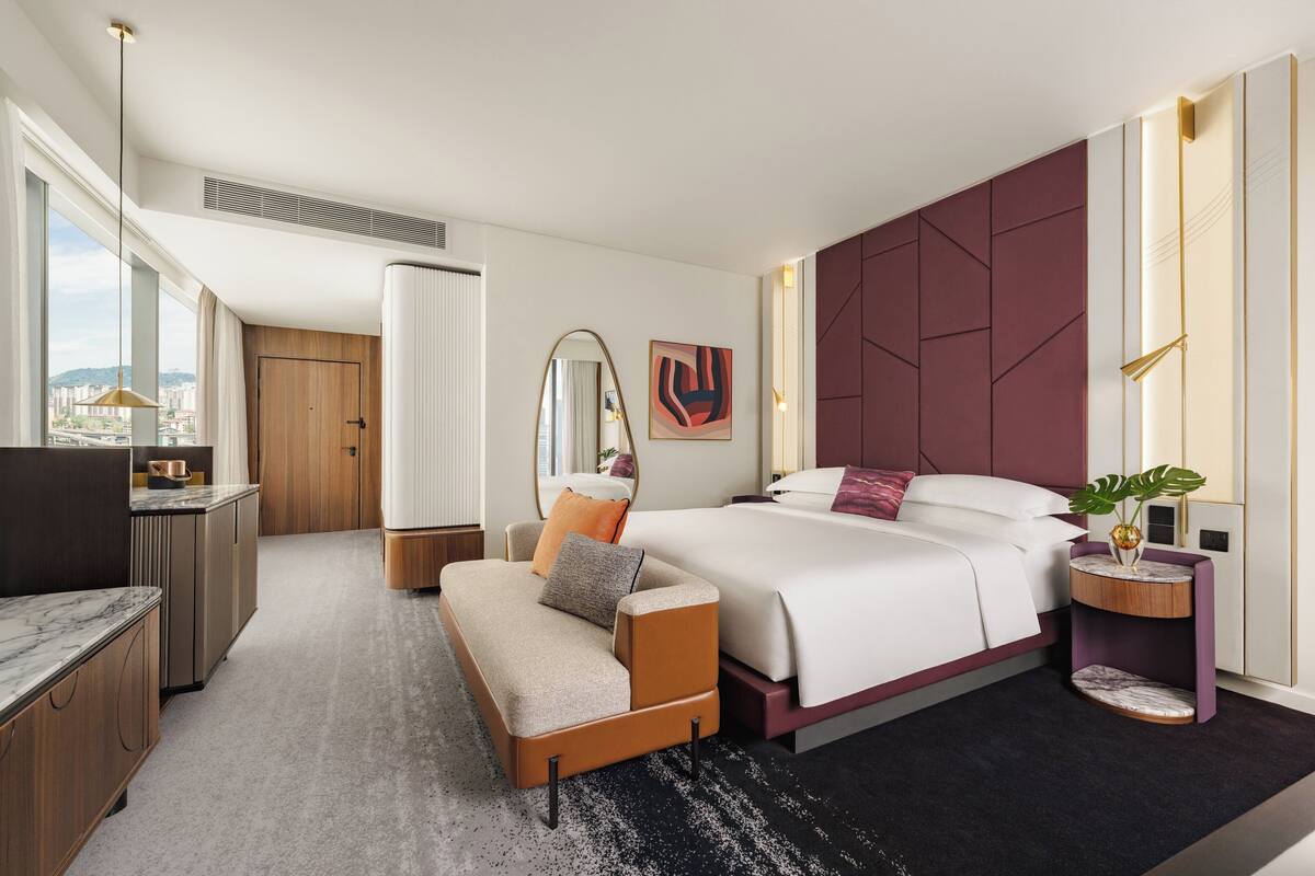 A modern hotel room with a large bed, a sofa, a desk, and a view of the city.