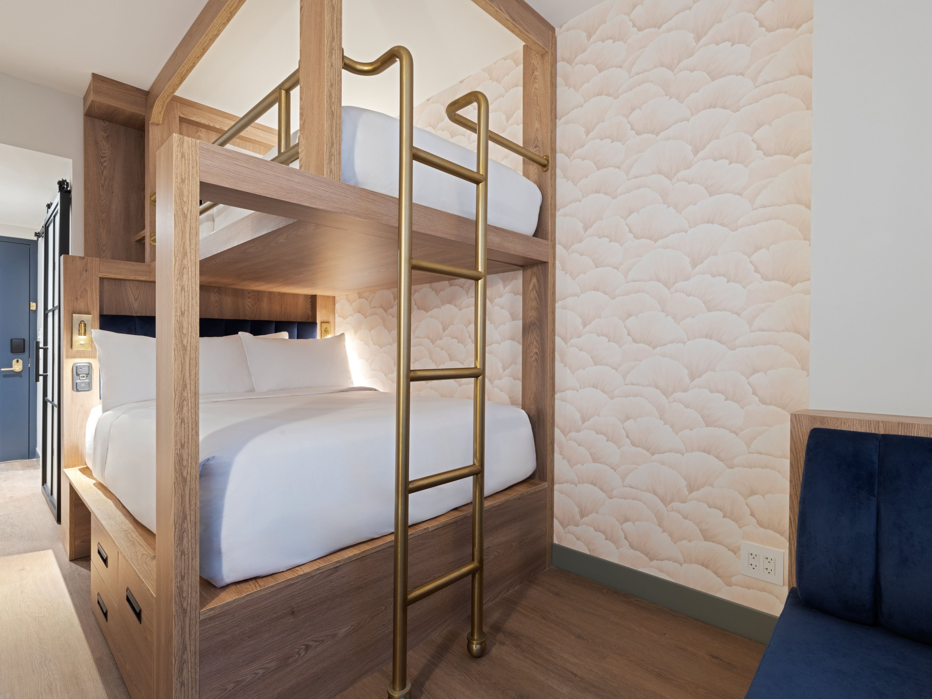Una cama litera con estructura de madera y escalera de latón, un sillón tapizado en azul y una pared con textura.