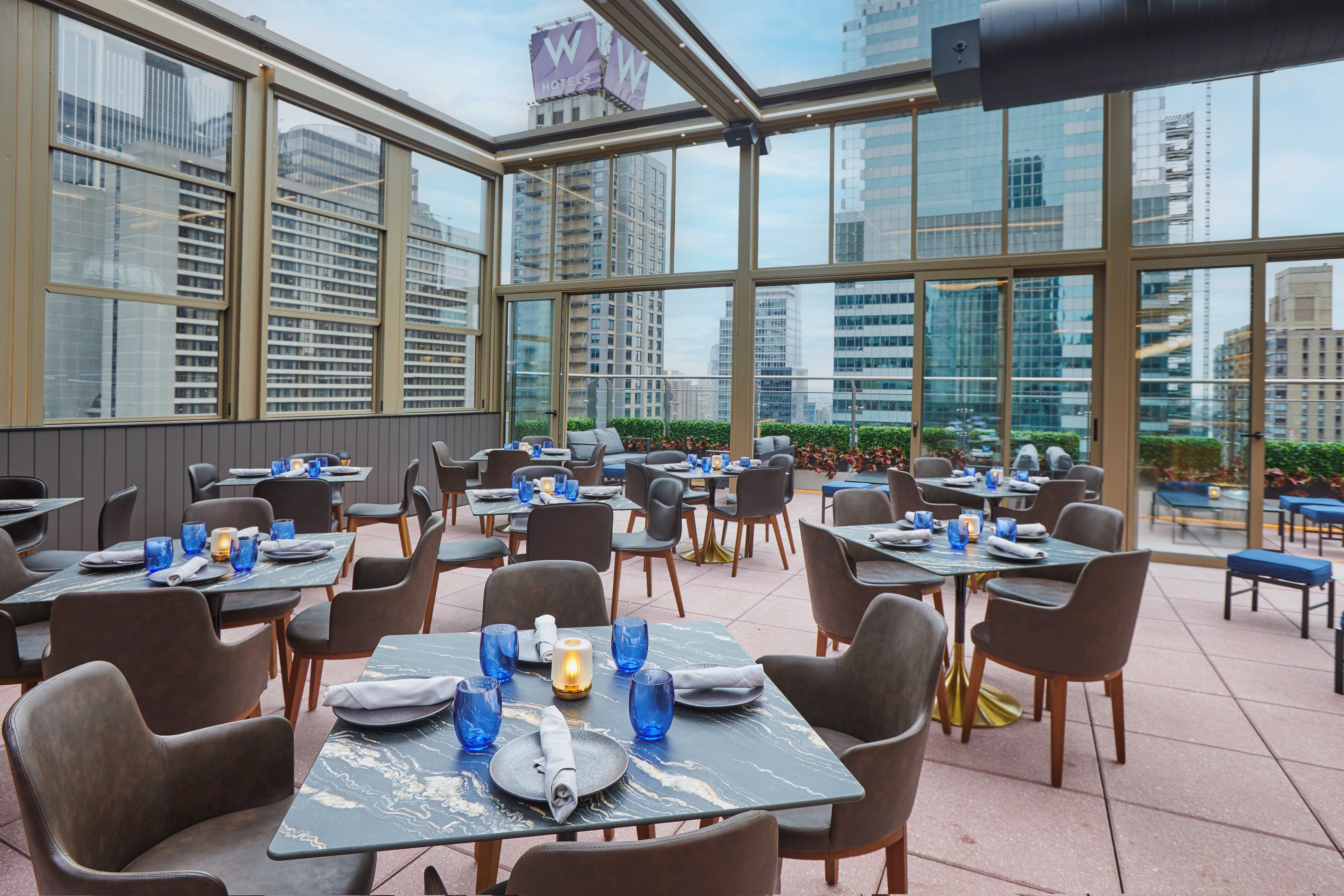 Un restaurant avec des tables dressées pour le service, de grandes fenêtres et une vue sur le paysage urbain.