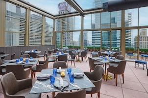 Um restaurante com mesas postas para refeições, grandes janelas e vista para o horizonte da cidade.