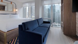 Una habitación de hotel moderna con cama, sofá, ventanas amplias, un televisor de pantalla plana y vista a la ciudad.
