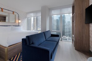 Una habitación de hotel moderna con cama, sofá, ventanas amplias, un televisor de pantalla plana y vista a la ciudad.