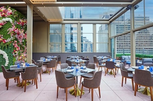 A restaurant with tables and chairs arranged for dining, large windows offering a view of the cityscape, and a decorative floral wall on one side.