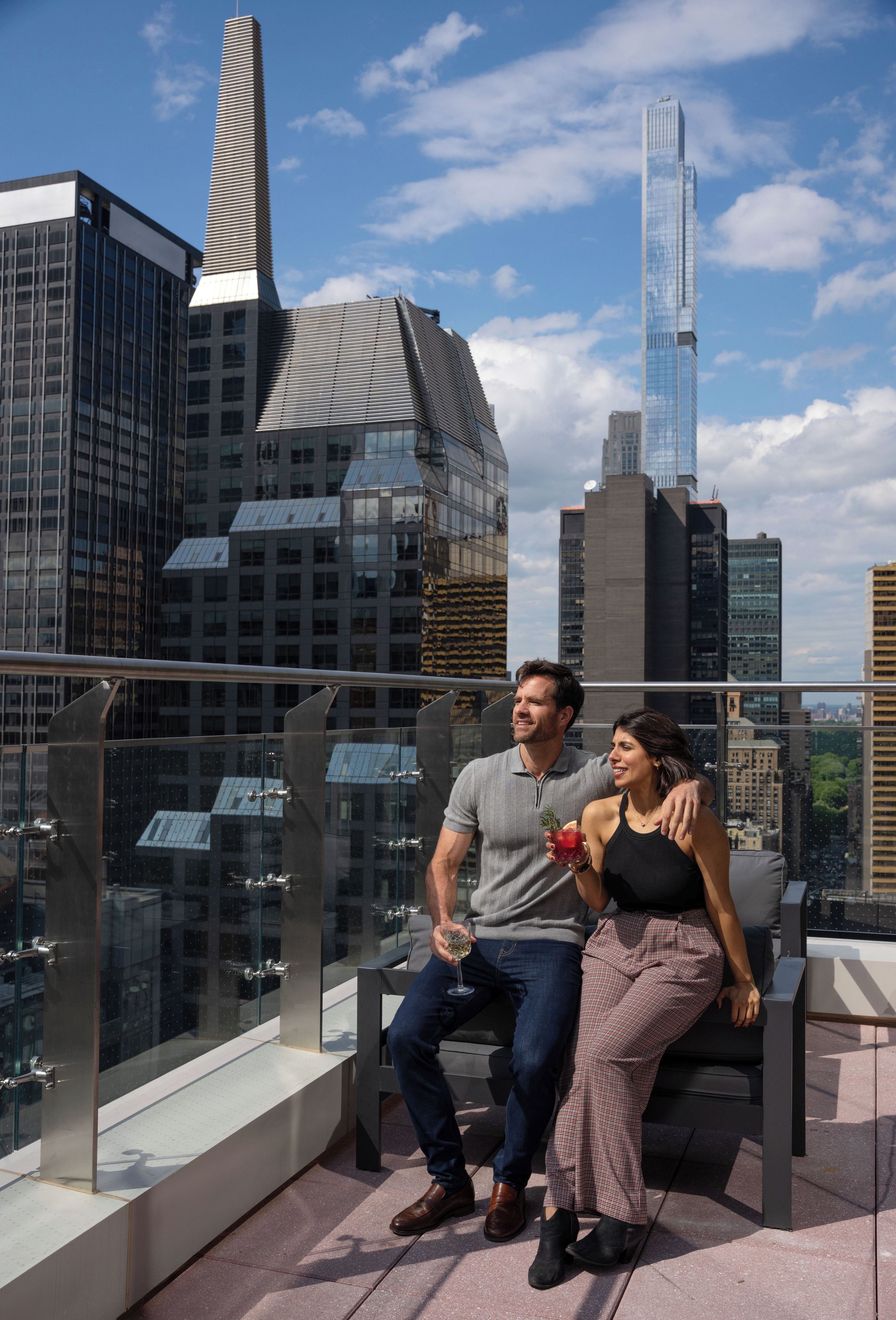 Una pareja sentada en un balcón con vista a un paisaje urbano, que incluye edificios modernos y un cielo despejado.