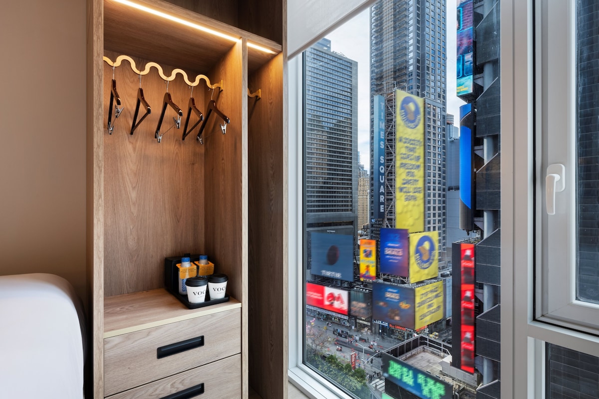 A hotel room with a wooden wardrobe, a window with a city view, and a small table with two containers.