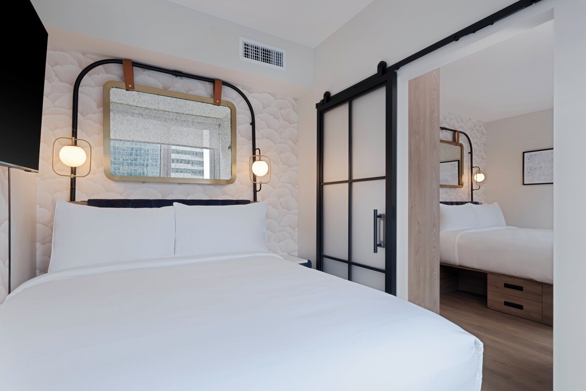 Una habitación de hotel moderna con una cama grande, un espejo montado en la pared y una puerta corredera que da a una cama más pequeña.
