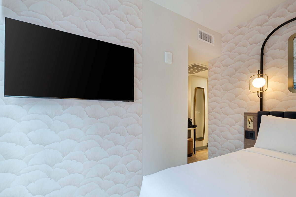 A hotel room with a large flat-screen TV mounted on the wall, a bedside table with a lamp, and a door leading to a bathroom.