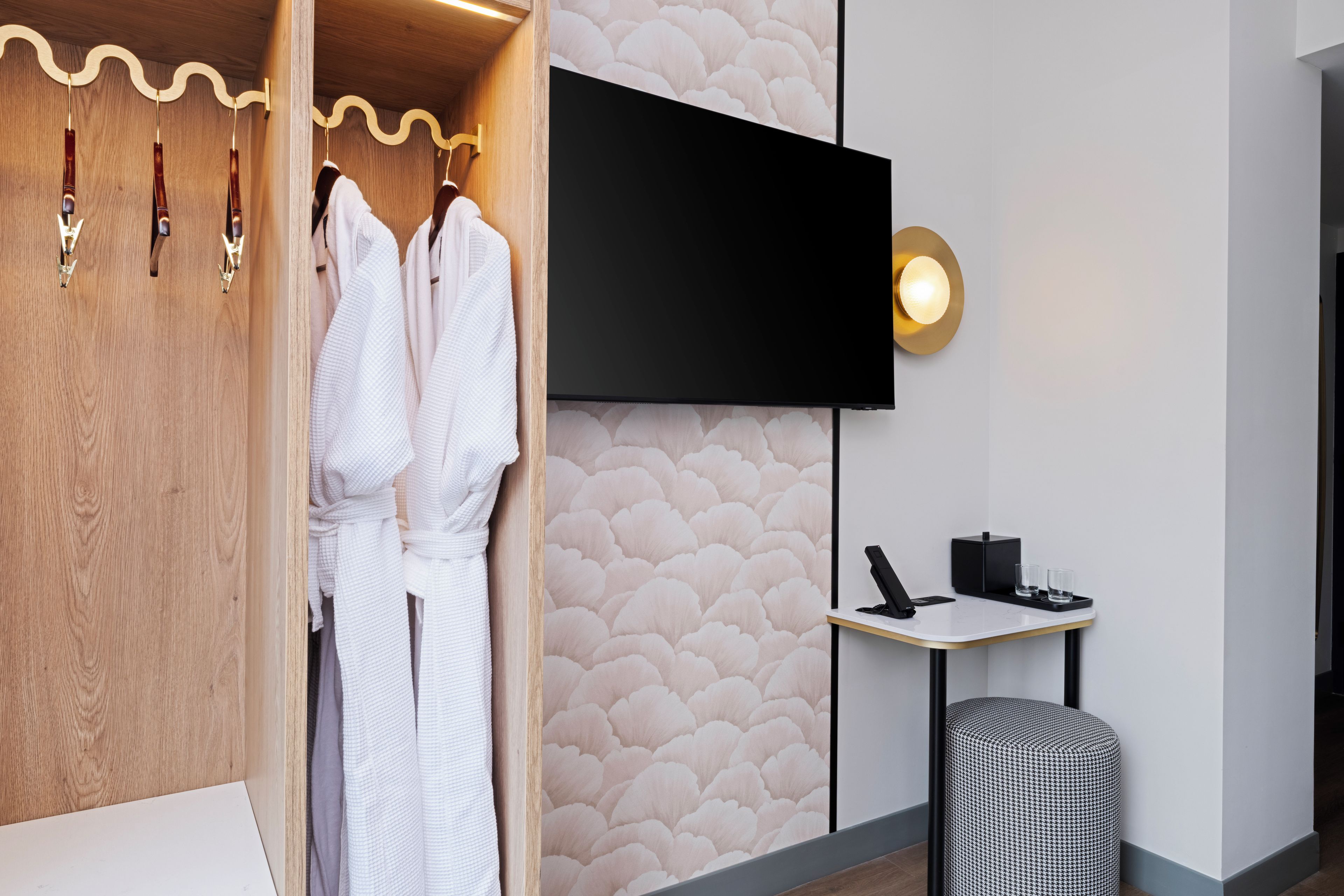 A modern bathroom with a wooden vanity, a flat-screen TV mounted on the wall, and a small table with a telephone and glasses.