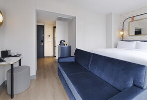 Quarto de hotel com uma cama, um sofá, uma mesinha com um telefone e copos, e uma lixeira.