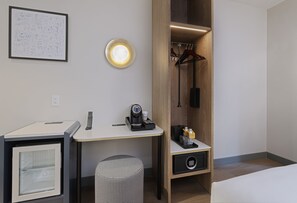 A hotel room with a desk, a coffee machine, a small cabinet, a hanging coat rack, and a bed. - voco Times Square - Broadway by IHG (New York)