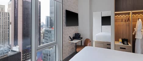 Ein Hotelzimmer mit großem Fenster, einem Bett, einem Schreibtisch mit Stuhl, einem Kleiderschrank und einem an der Wand befestigten Fernseher.