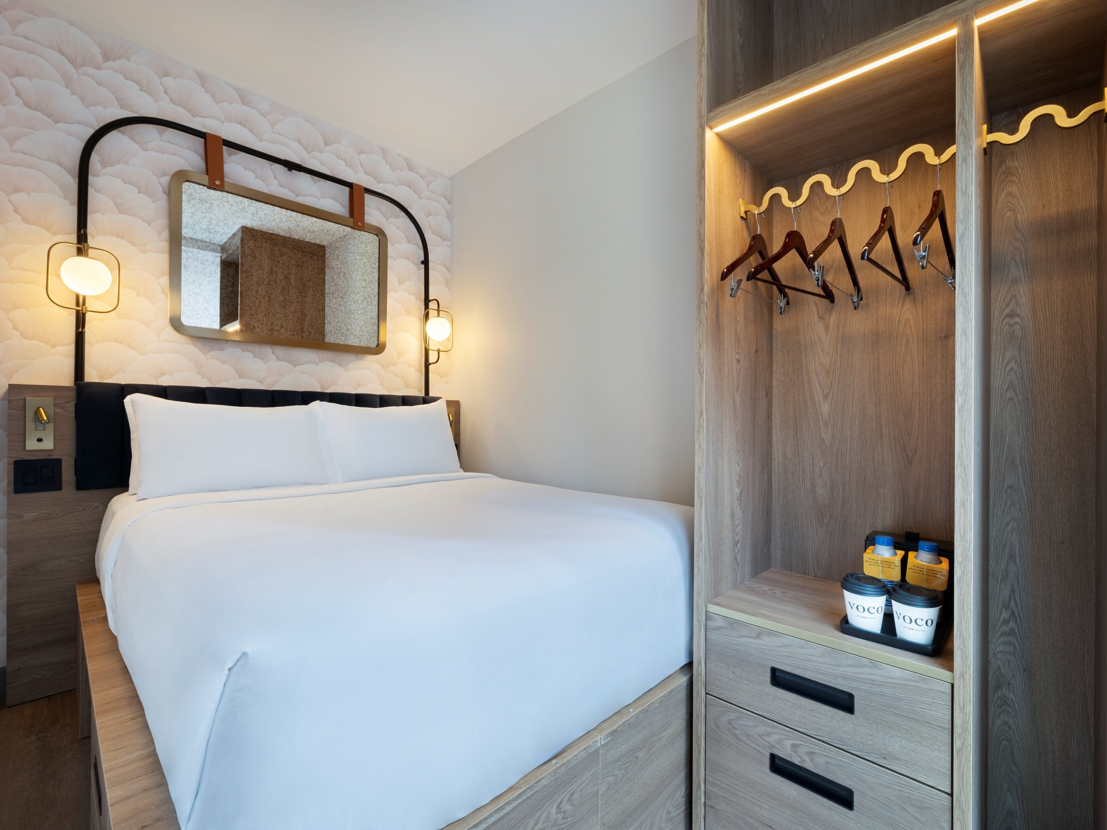 Una cama bien hecha con ropa blanca, un espejo montado en la pared y un armario de madera con espacio para colgar.