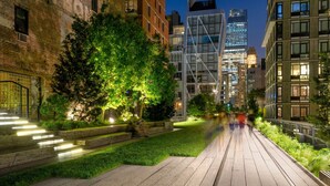 Un camino urbano nocturno con edificios modernos, un árbol bien iluminado y un muro de ladrillo.