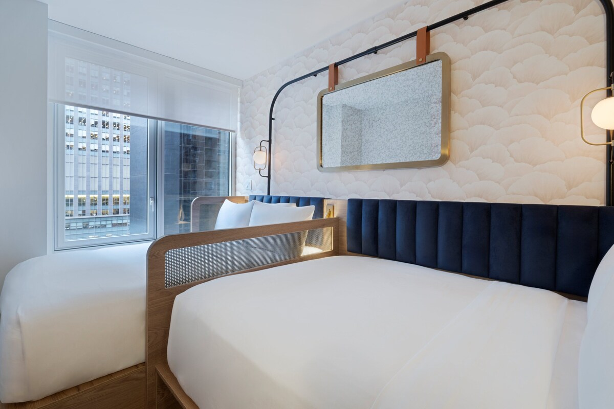 Habitación de hotel con una cama grande, una cama más pequeña, una ventana con vistas a la ciudad, un espejo en la pared y una lámpara de pared.