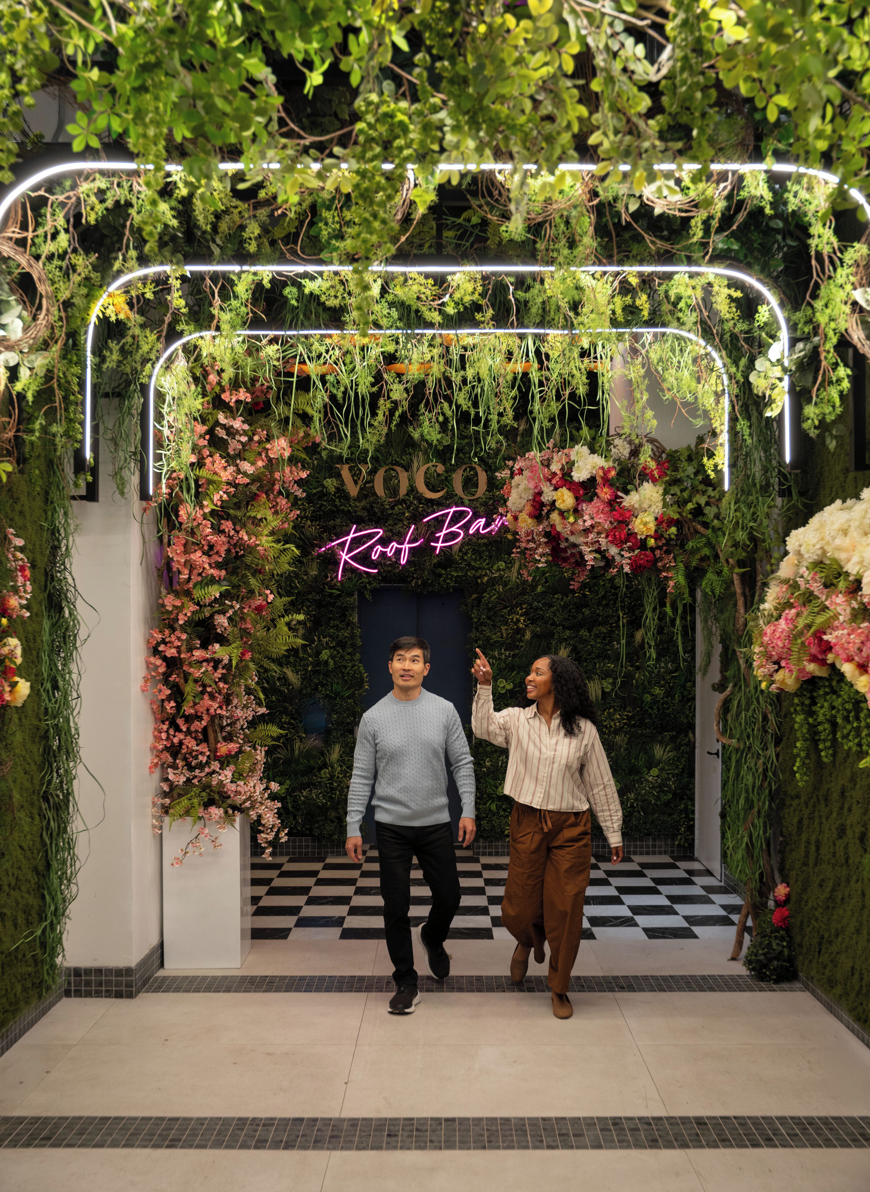 Una coppia cammina attraverso una porta adornata da un cartello luminoso con scritto 'Voco Roof Bar', immersa in una lussureggiante vegetazione e decorazioni floreali.