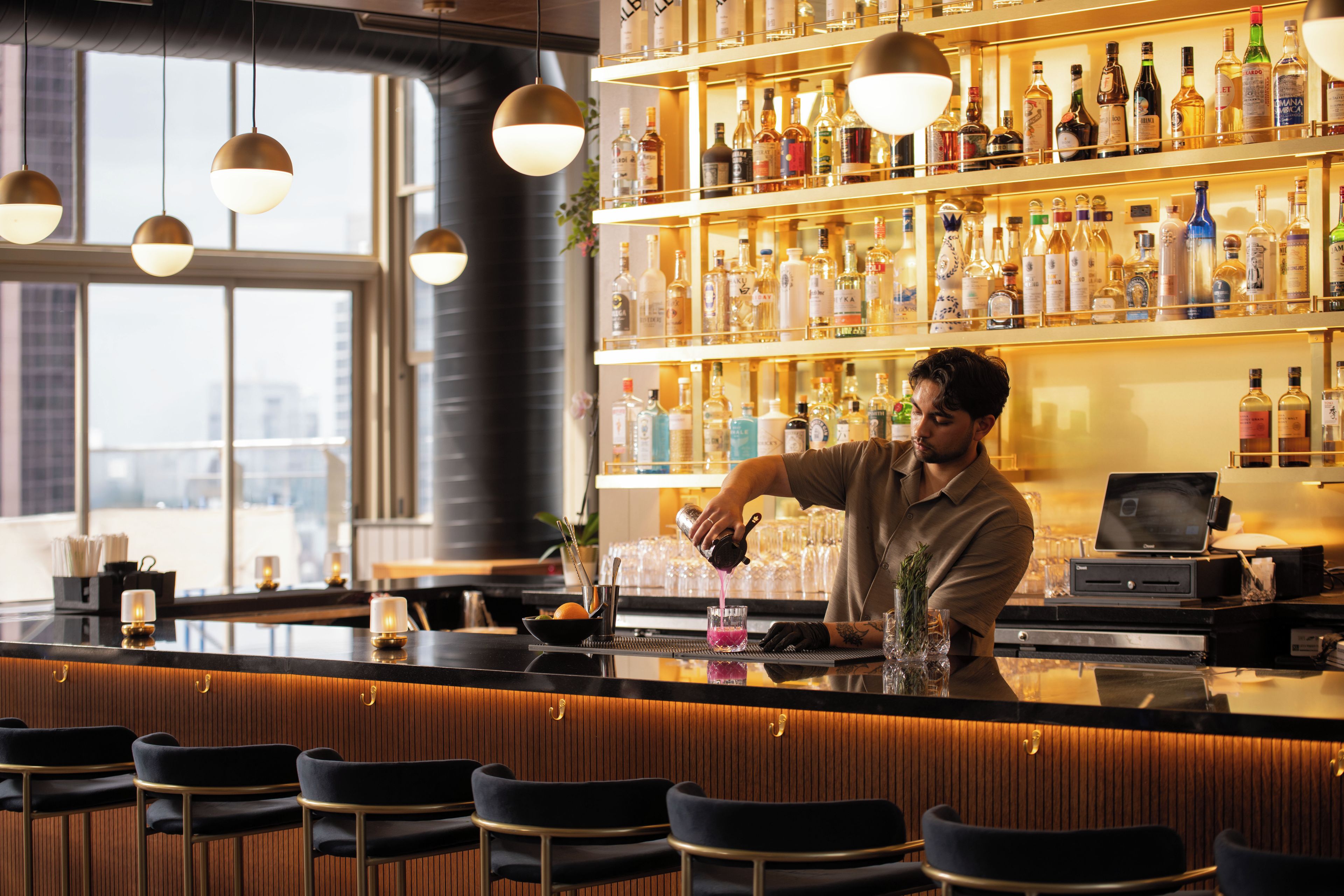 A bar area with a bartender preparing a drink, shelves of liquor bottles, and bar stools.