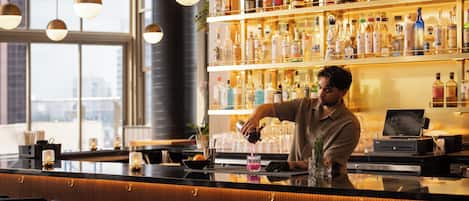 A bar area with a bartender preparing a drink, shelves of liquor bottles, and bar stools.