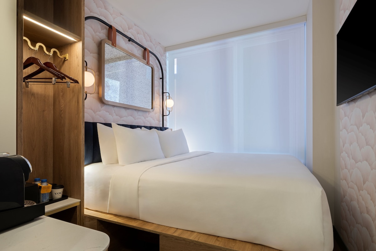 A modern hotel room with a large bed, a wooden desk, a mirror, and a TV mounted on the wall.