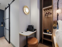 Quarto de hotel moderno com porta deslizante, pequena mesa com cafeteira, banqueta de couro marrom, guarda-roupa de madeira com espelho e cama branca.