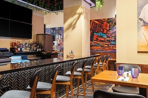 Zona de bar con una barra larga, diversas opciones de asientos que incluyen taburetes y mesas, y un mural con vistas a la ciudad.