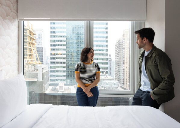 A hotel room with a large window, a bed, and a view of city buildings. - voco Times Square - Broadway by IHG (New York)