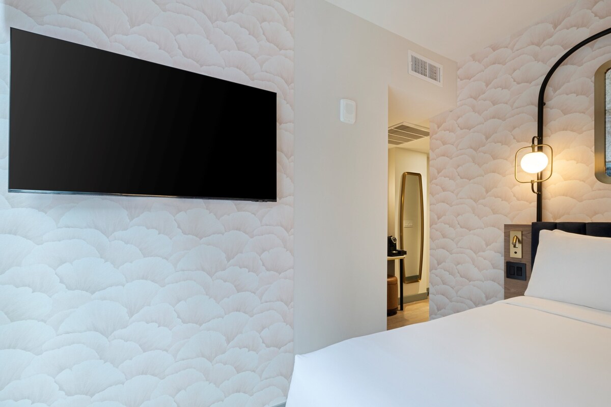 Habitación de hotel con un televisor de pantalla plana grande montado en la pared, una mesita de noche con lámpara y una puerta que da al baño.