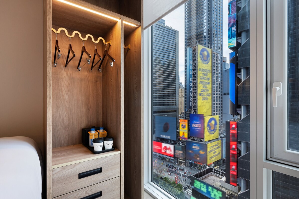 A hotel room with a wooden wardrobe, a window with a city view, and a small table with two containers.