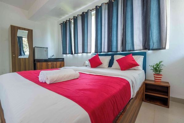 Una cama bien tendida con ropa de cama roja y blanca, una mesita de noche con una planta en maceta y un espejo en la pared.