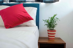 Una cama con un almohadón rojo y una maceta a rayas sobre una mesita de noche de madera.
