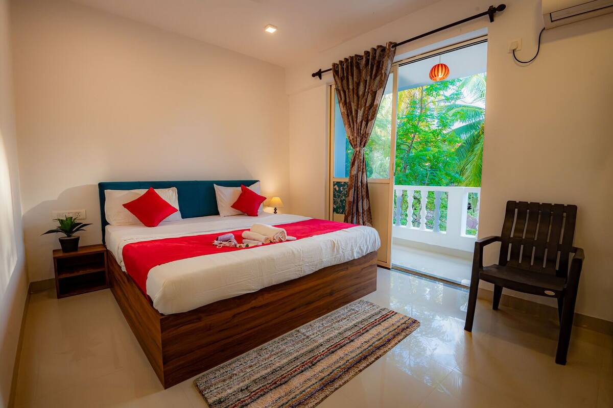 A bedroom with a wooden bed, red and white bedding, a small bedside table with a plant, and a view of greenery through a balcony door.