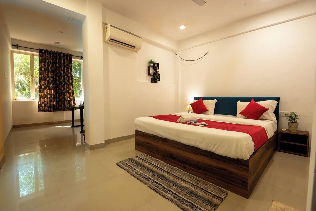 A modern bedroom with a large bed, a bedside table, an air conditioning unit, and a view of the outdoors.