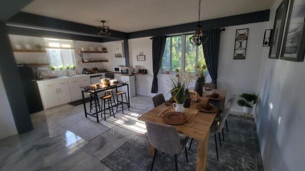 A modern kitchen with white cabinets, a dining area with a wooden table and chairs, and a large window with curtains. - Visconti Guest House (Cottage)