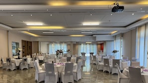 Une salle de banquet spacieuse, dotée de nombreuses tables rondes, chacune recouverte d’une nappe blanche et entourée de chaises.