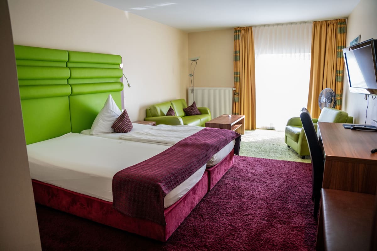 A hotel room with a green headboard bed, a second bed with a red bedspread, a green sofa, a wooden desk, and a television.