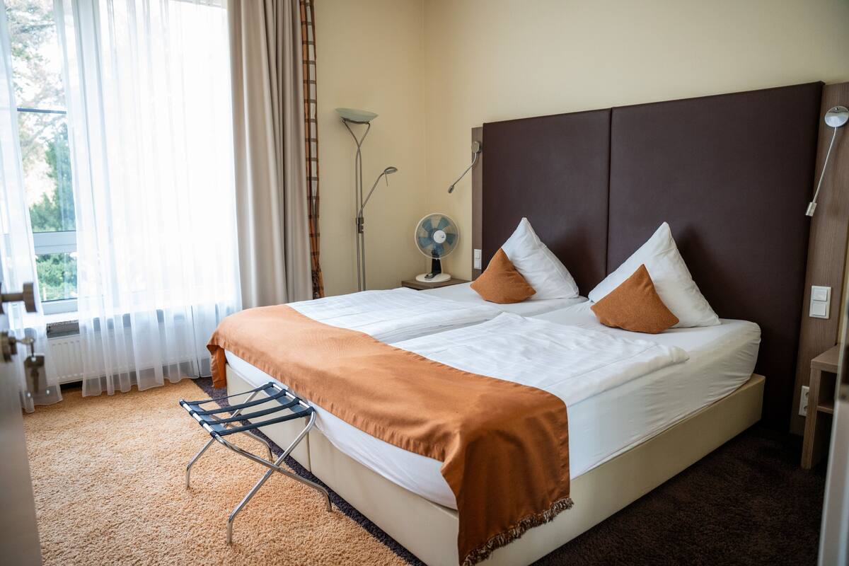 A hotel room with a large bed, a standing fan, a nightstand, and a window with curtains.