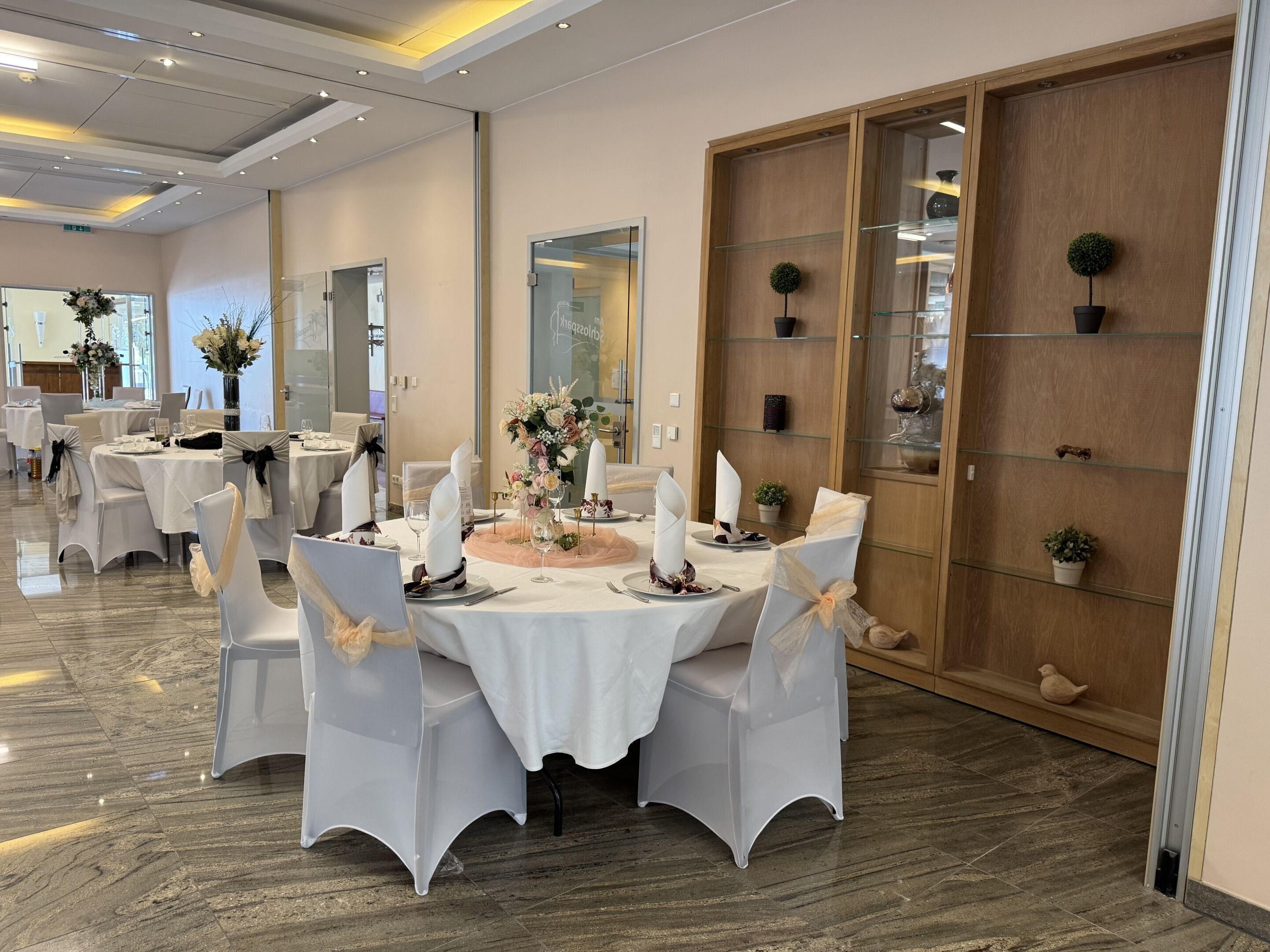 Un salón de banquetes con mesas redondas dispuestas para un evento formal, que incluye manteles blancos, sillas con fundas blancas y centros de mesa florales.