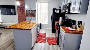 Una cocina moderna con armarios grises, encimera de madera y una isla central. Hay un televisor montado en la pared, una pared de acento de ladrillo y una ventana con persianas.