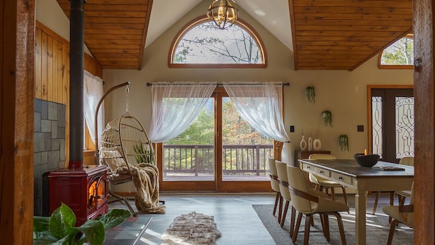 A cozy room with a stone fireplace, a hanging chair, a dining table with chairs, and a view of the outdoors through large windows.
