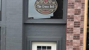 A building with a white door, a sign above it reading "The Cross Bell," and a small outdoor area with a doormat and a utility box. - The Cross Bell Downtown 201 (Pawhuska)