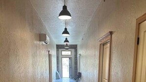 A long, narrow hallway with wooden flooring and white walls. There are two black pendant lights hanging from the ceiling, and a door on the right side.