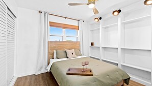 A bedroom with a wooden headboard, a bed with a green bedspread, a wooden tray with two glasses, and a ceiling fan.