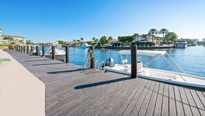A wooden dock with boats, a clear blue sky, and a marina with buildings and palm trees in the background.