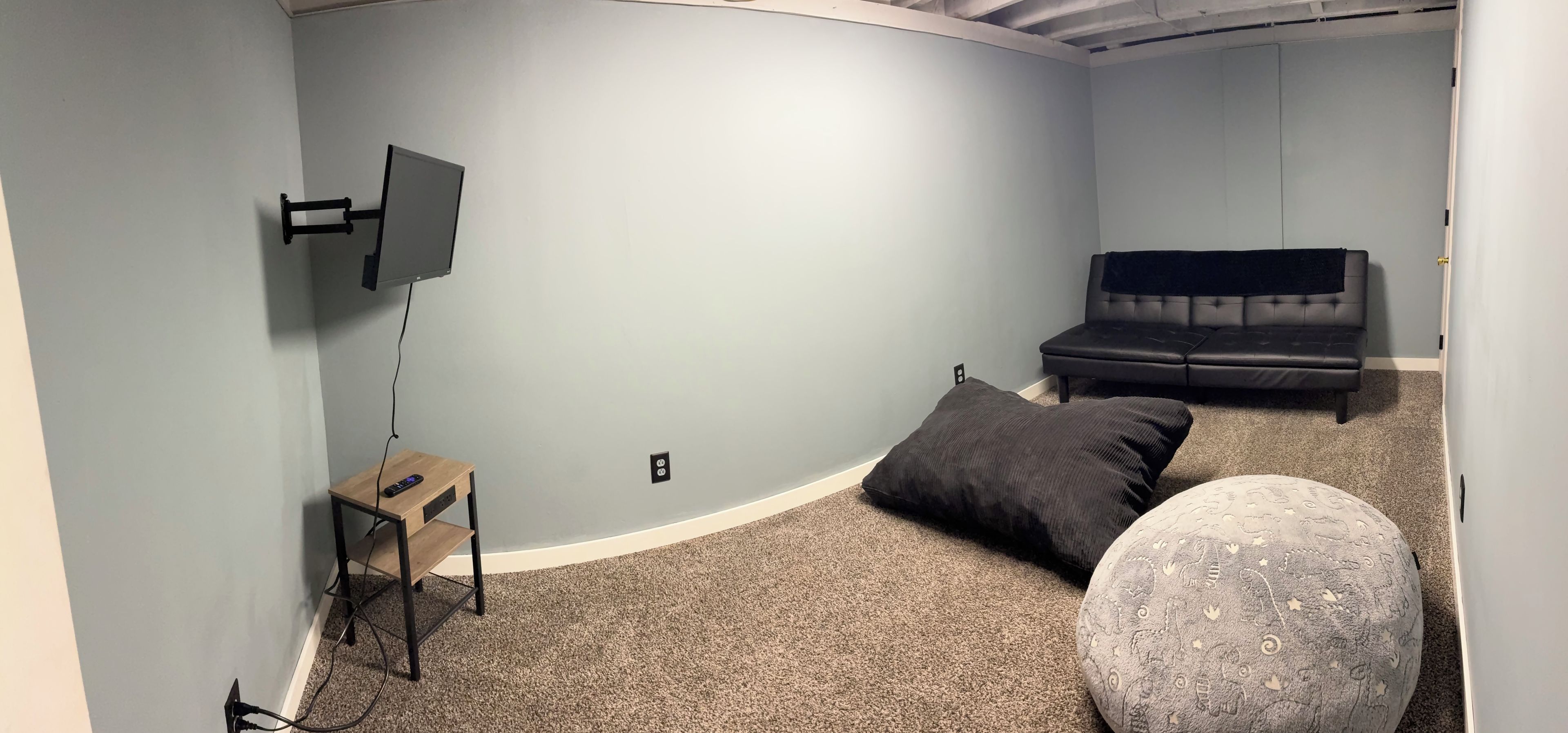A room with a flat-screen TV mounted on the wall, a small wooden side table with a remote control, a black couch, and a round ottoman with a patterned design.