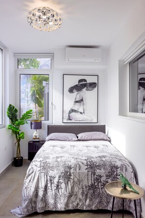 A bedroom with a bed, a side table, a potted plant, a wall-mounted air conditioner, and a window with a view of greenery. - Villa in Kournas with private pool and stunning sea views (Kournas)